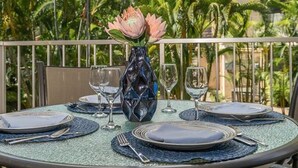 A round table set for dining with a vase of flowers, surrounded by chairs, with a view of palm trees and a building in the background.