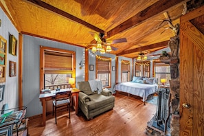 A cozy room with a wooden ceiling, a bed with a blue bedspread, a desk with a chair, and a window with blinds.