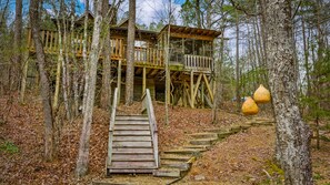 A wooden cabin with a wooden staircase leading to a deck, surrounded by trees and a forest floor covered with leaves.