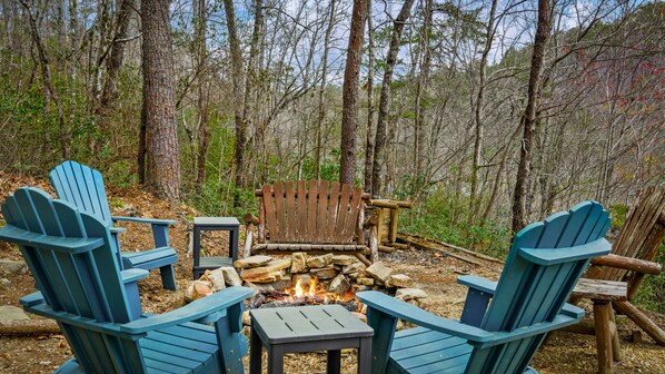 A campfire surrounded by Adirondack chairs and a wooden bench in a forest setting.