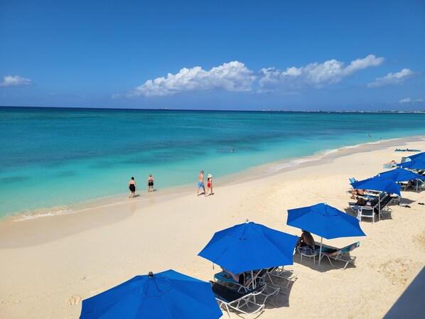 Une plage à l’eau turquoise et limpide, avec un rivage sablonneux et plusieurs parasols bleus qui offrent de l’ombre.