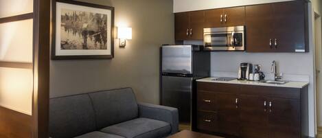 A modern hotel room with a sofa, a coffee table, a kitchen area with a refrigerator, microwave, and cabinets.
