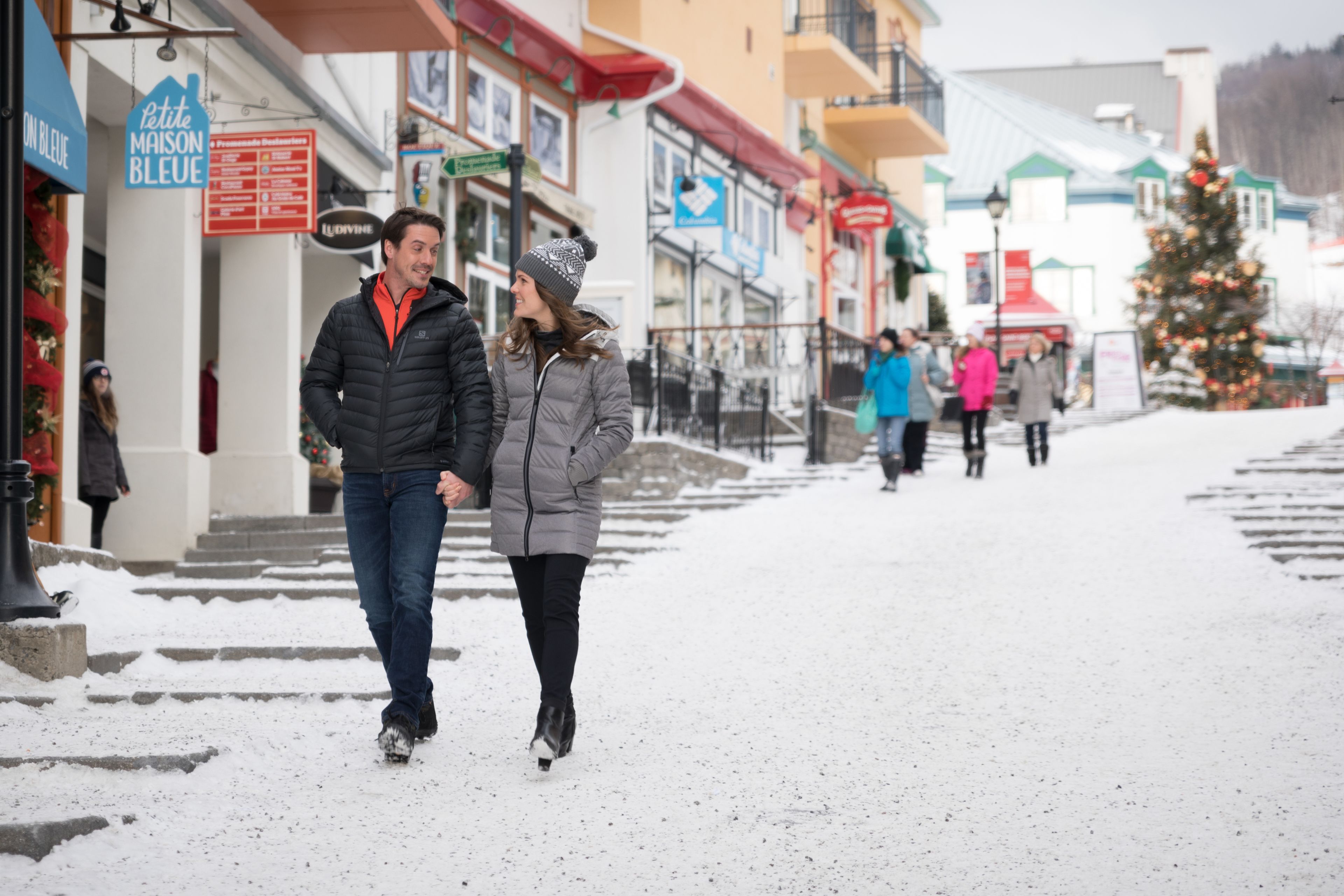 A couple walking on a snow-covered street with buildings and a Christmas tree in the background.