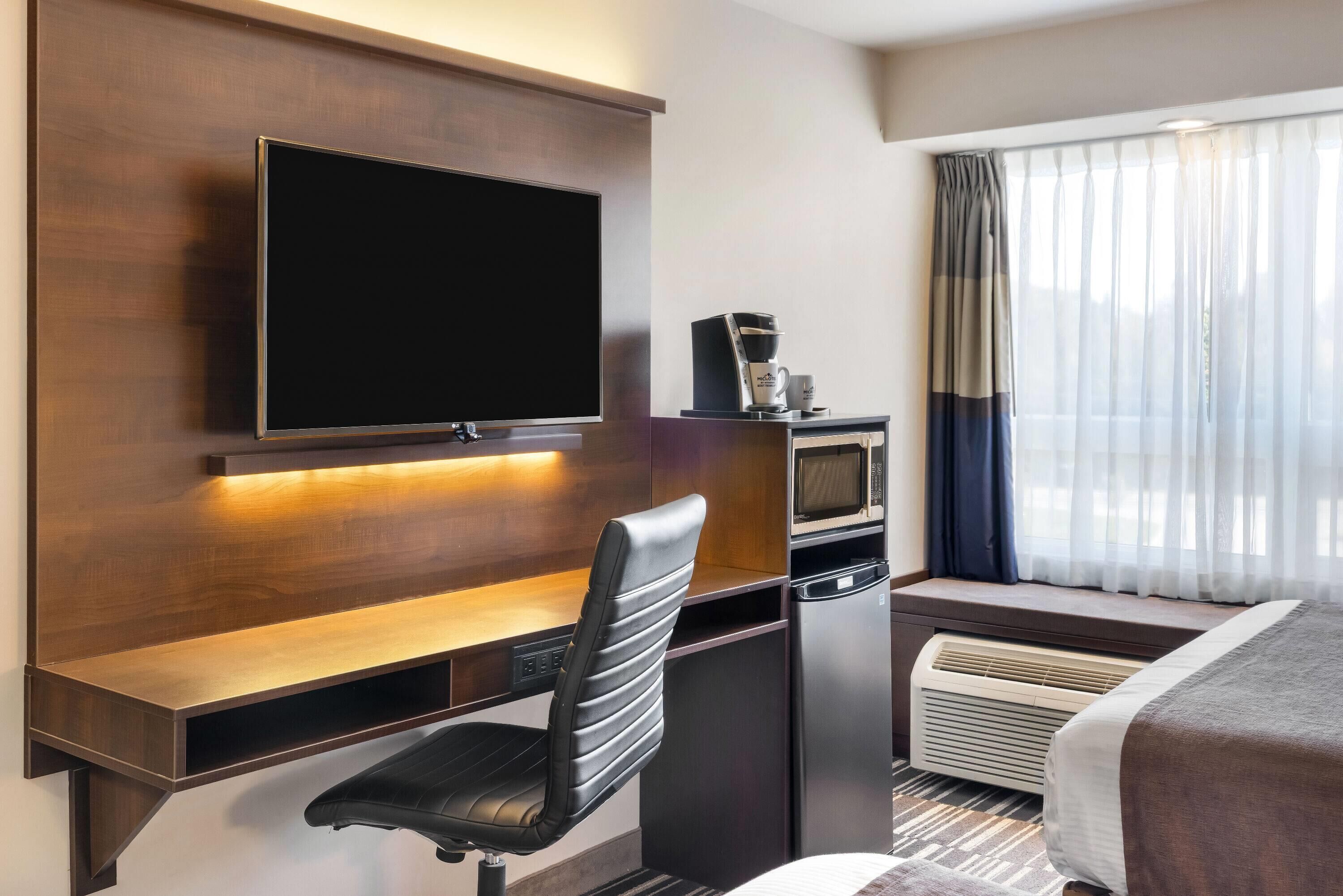 Habitación de hotel con una televisión de pantalla plana montada en una pared de madera, un escritorio con una silla, un microondas, una cafetera y una cama con ventana.