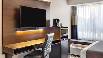 Habitación de hotel con una televisión de pantalla plana montada en una pared de madera, un escritorio con una silla, un microondas, una cafetera y una cama con ventana.