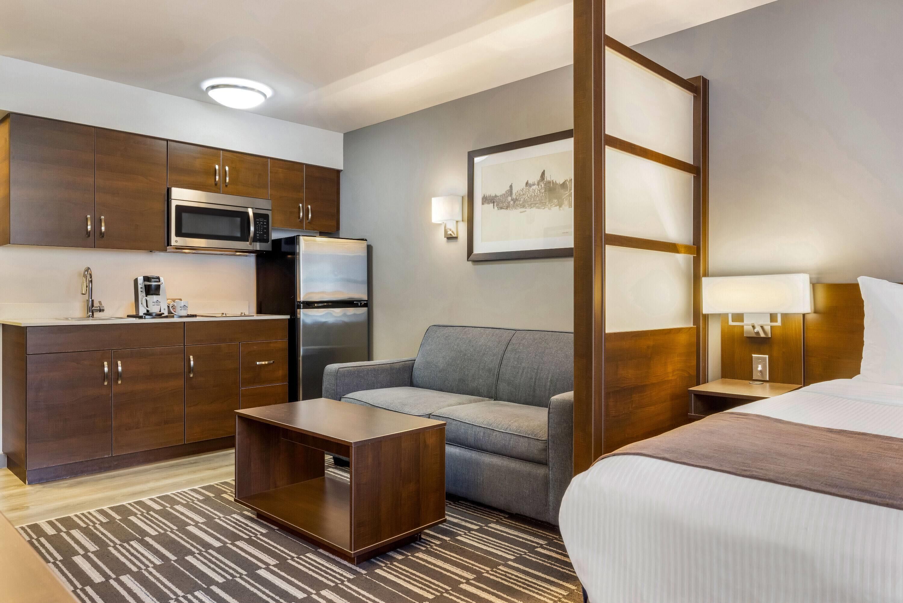 A compact hotel room with a kitchenette, a gray sofa, a wooden coffee table, a bed, and a framed picture on the wall.