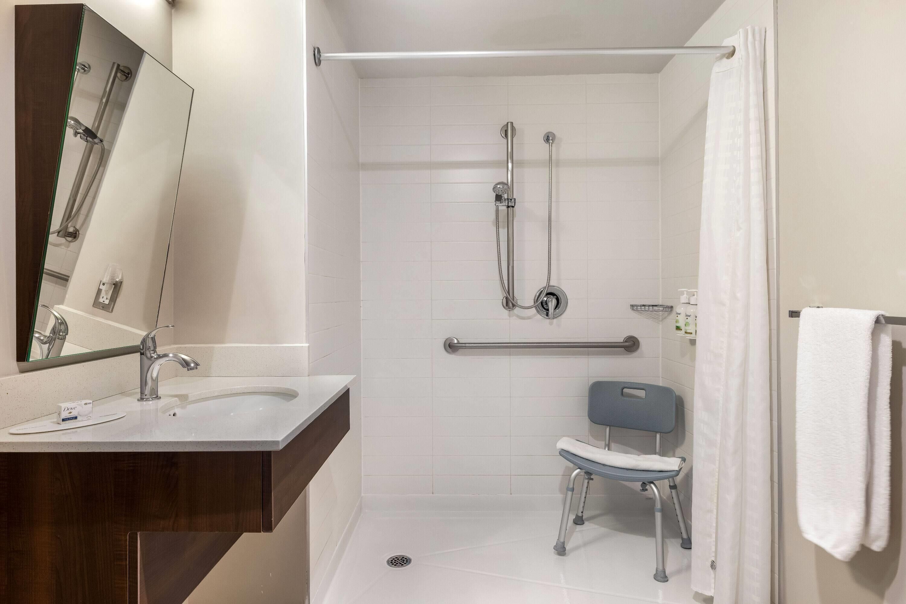 A modern bathroom with a white sink, a mirror, a shower with grab bars, and a wheelchair-accessible bench.