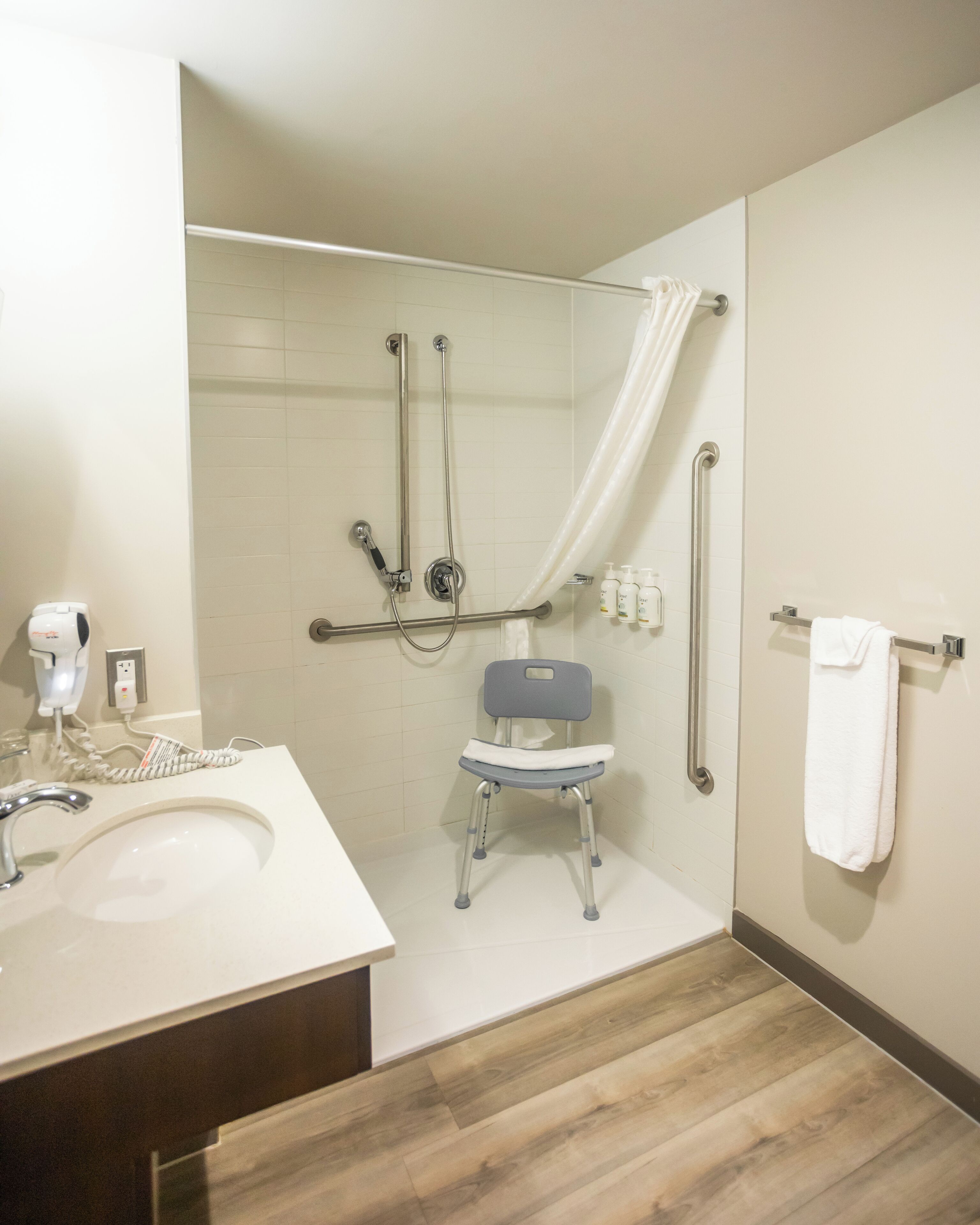 Een moderne badkamer met een douche, handgrepen, een rolstoeltoegankelijke bank, een wastafel en een handdoekhouder.
