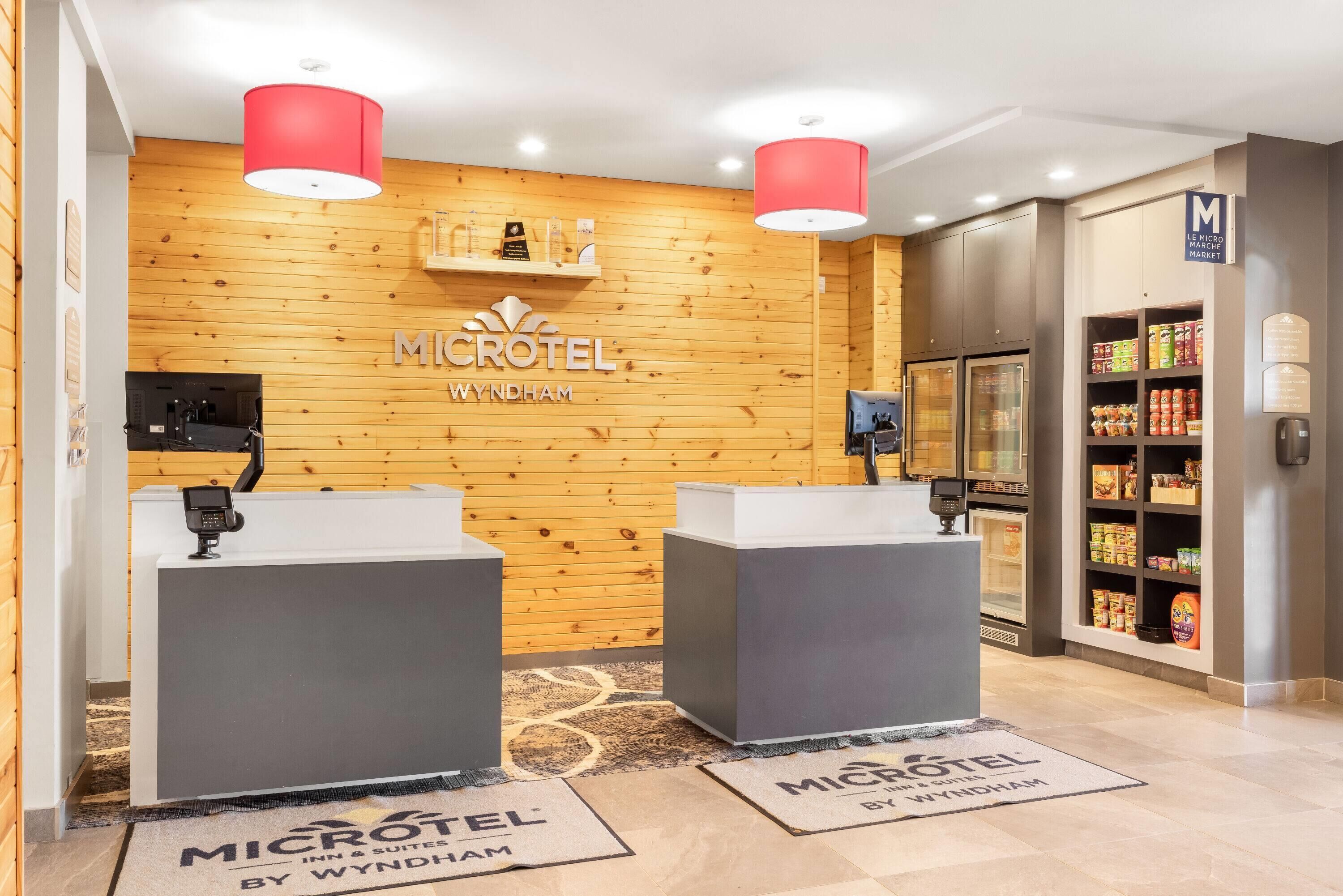 A reception area with a wooden wall, two reception desks, a sign for 'MICROTEL WYNDBHAM', and a refrigerator stocked with beverages.