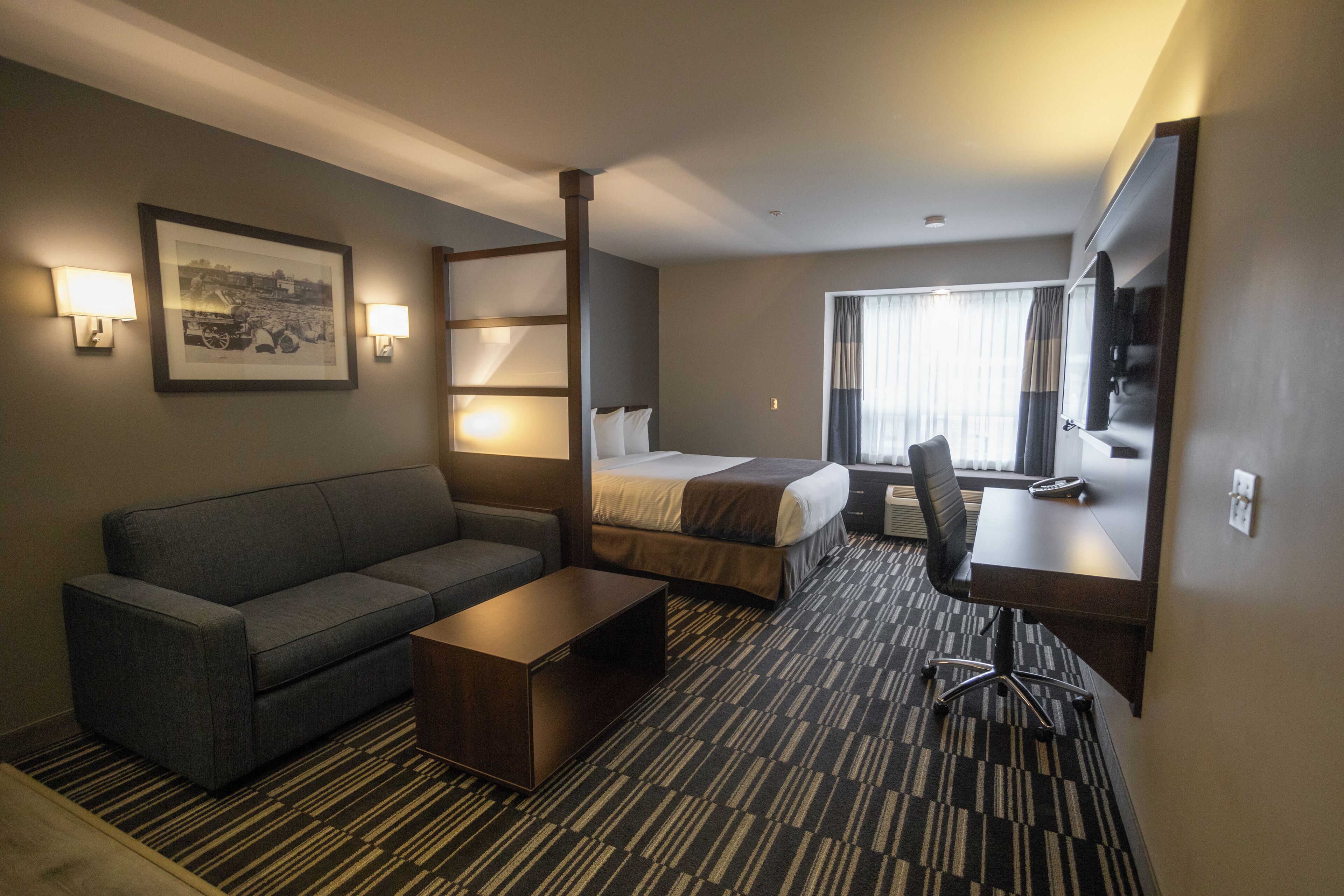 A hotel room with a sofa, a bed, a desk, and a chair. There are two wall-mounted lamps and a framed picture on the wall.