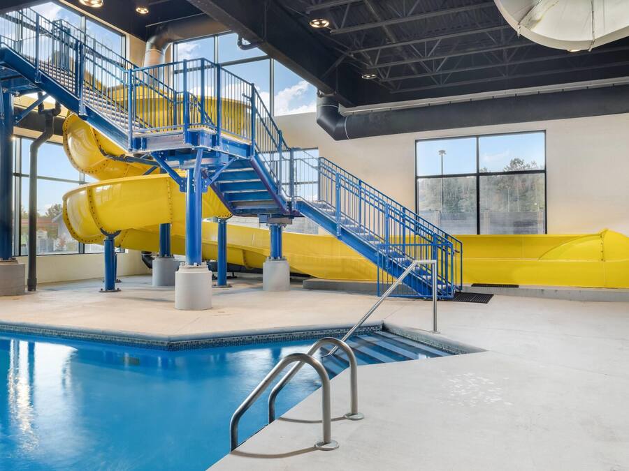 An indoor swimming pool with a blue and yellow water slide, stairs, and a view of the outside through large windows.