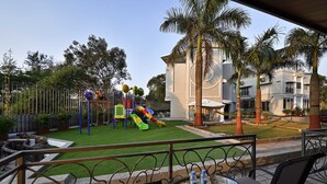 A playground with slides and swings, surrounded by palm trees and a building with balconies. - Meritas Countryside Resort - Lonavala (Mawal)