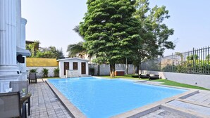 A swimming pool with a tiled deck, a small building with a door, and a large tree. - Meritas Countryside Resort - Lonavala (Mawal)