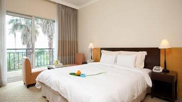 A hotel room with a large bed, two bedside tables, a chair, a balcony with curtains, and a view of palm trees.