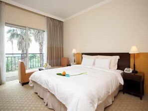 A hotel room with a large bed, two bedside tables, a chair, a balcony with curtains, and a view of palm trees. - Suites Hotel Jeju (Seogwipo)