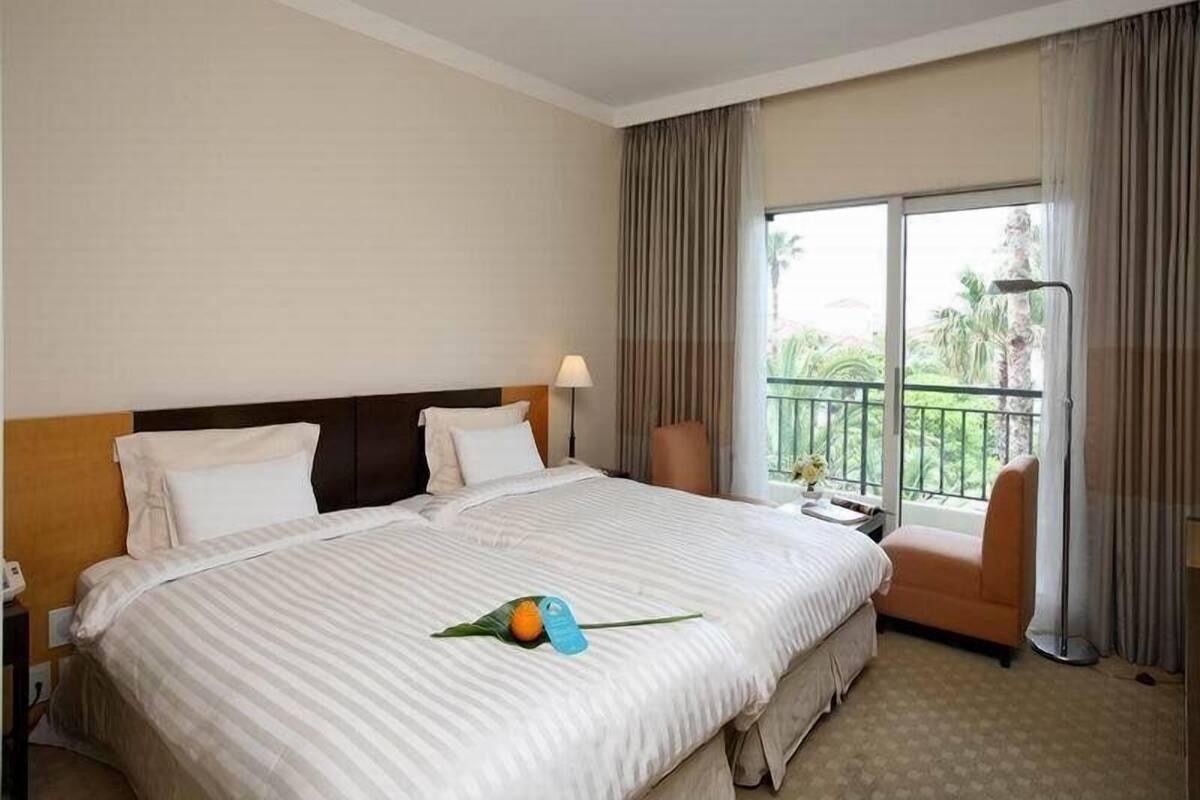 A hotel room with a large bed, a chair, a nightstand, and a window with a view of greenery.