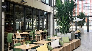 An outdoor seating area with wooden tables and chairs, a long bench with cushions, and large potted plants. - Sudima Christchurch City (Christchurch)