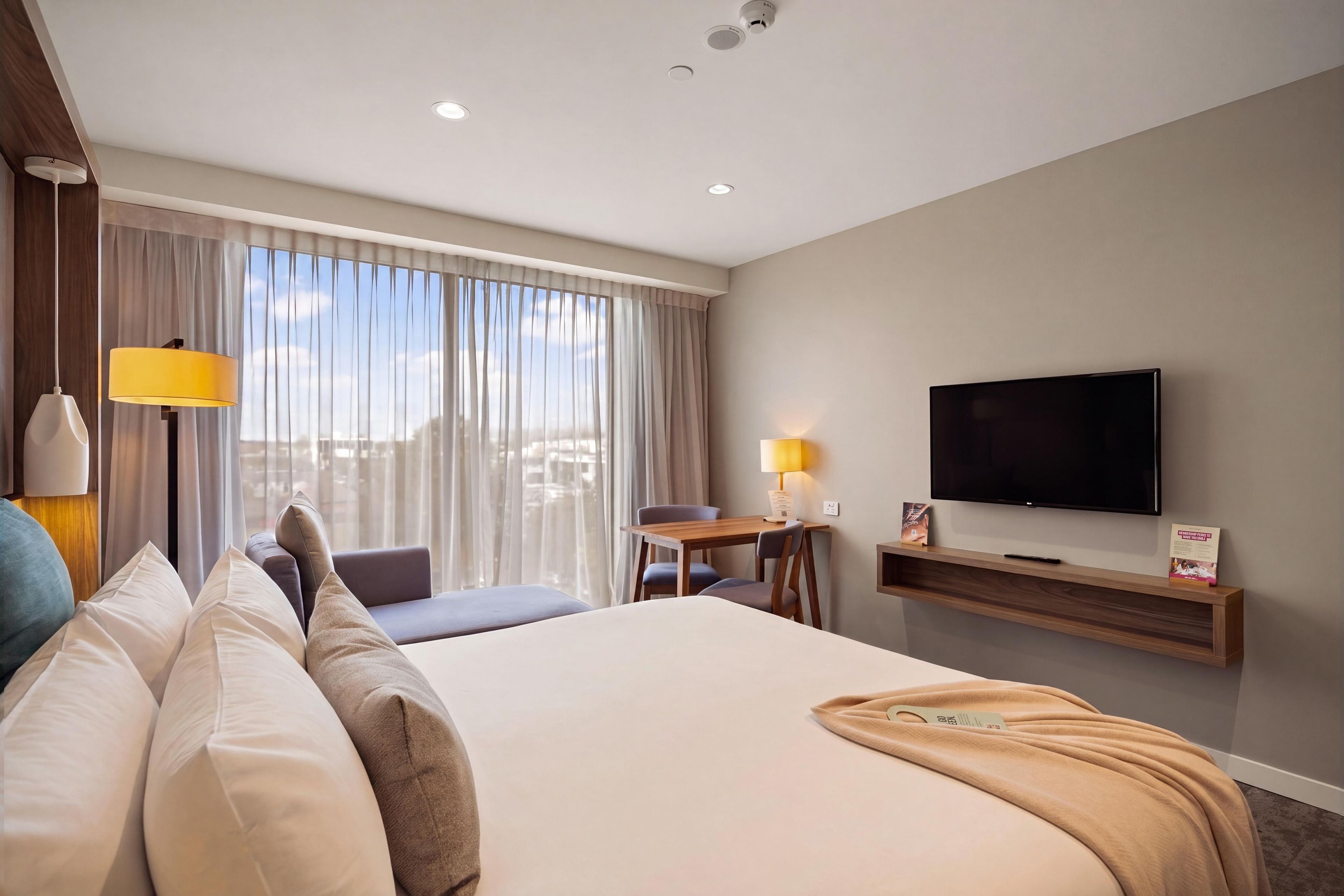 A hotel room with a large bed, a desk with a lamp, a television, and a window with curtains.