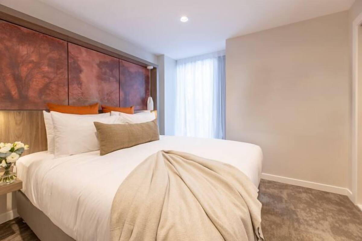 A hotel room with a large bed, a wooden headboard, a nightstand with a vase of flowers, and a window with curtains.