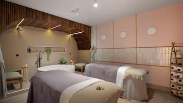 A modern spa room with a large bathtub, two treatment beds, a wooden ceiling, and a wall-mounted shelf with towels.