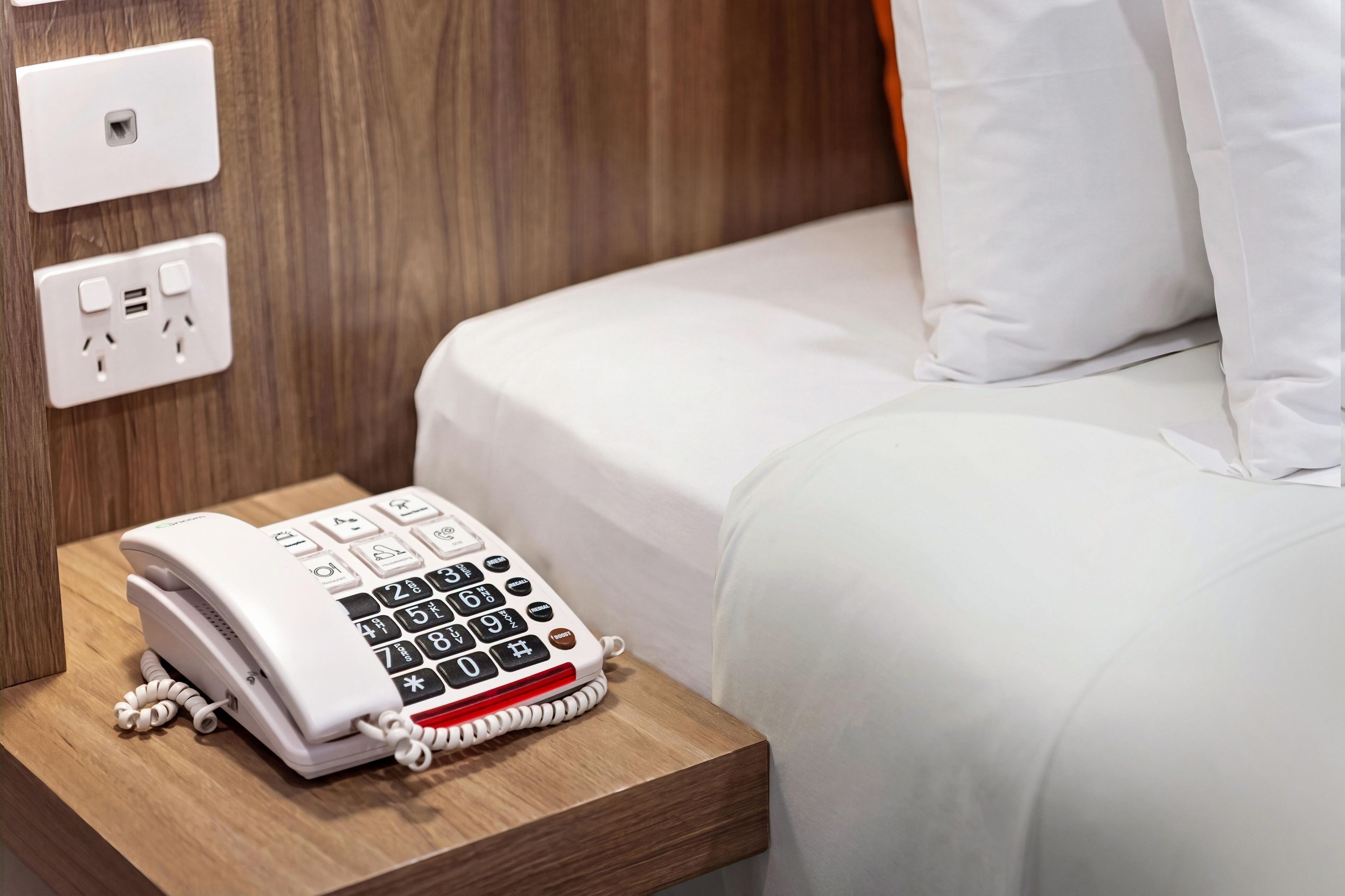 A hotel room with a bedside telephone, a power outlet, and a wall socket.