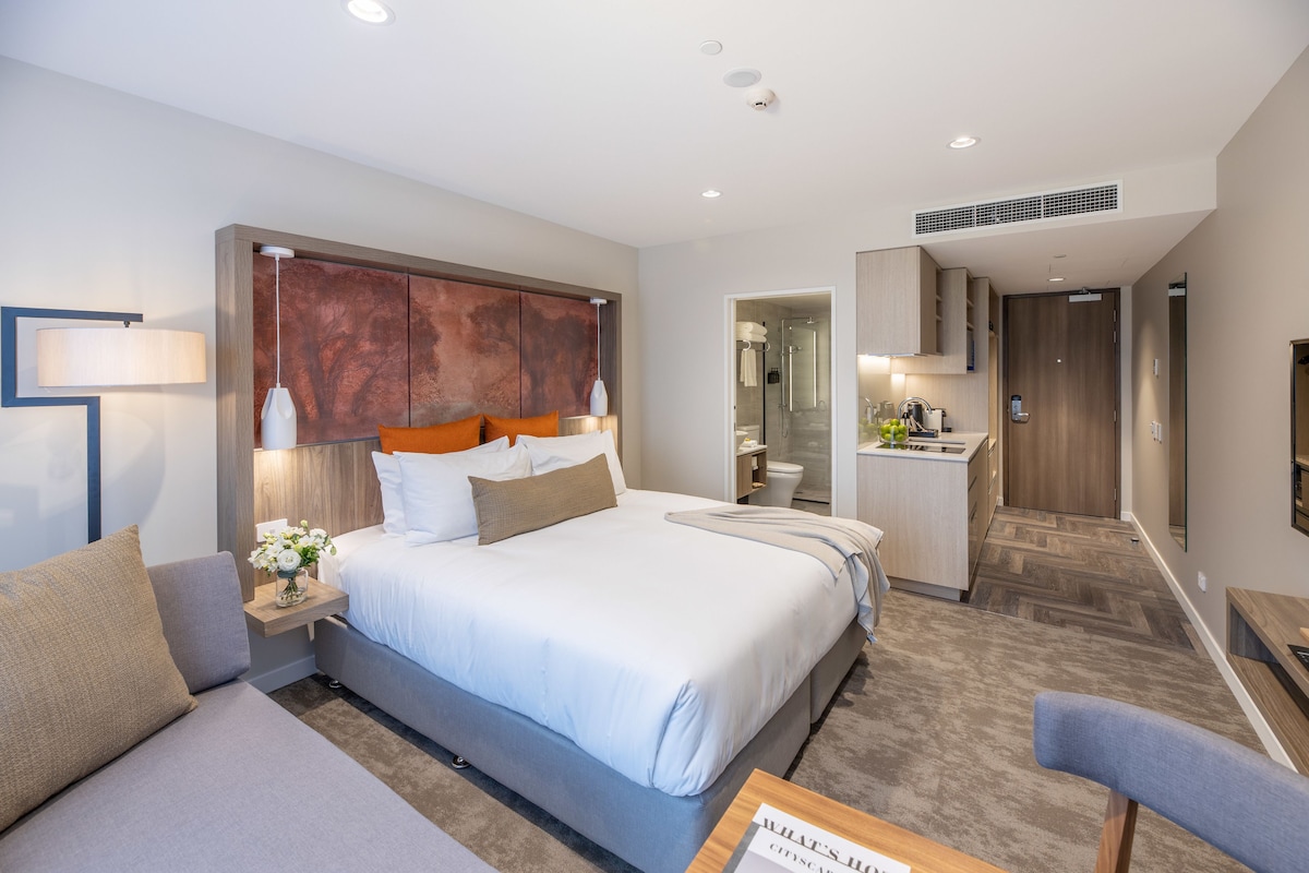 Una habitación de hotel moderna con una cama grande, zona de estar, kitchenette y baño.