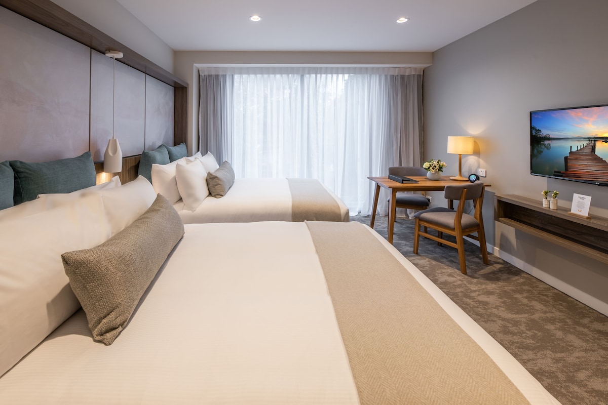 Habitación de hotel con dos camas, un escritorio con lámpara, televisión y vista a un muelle.