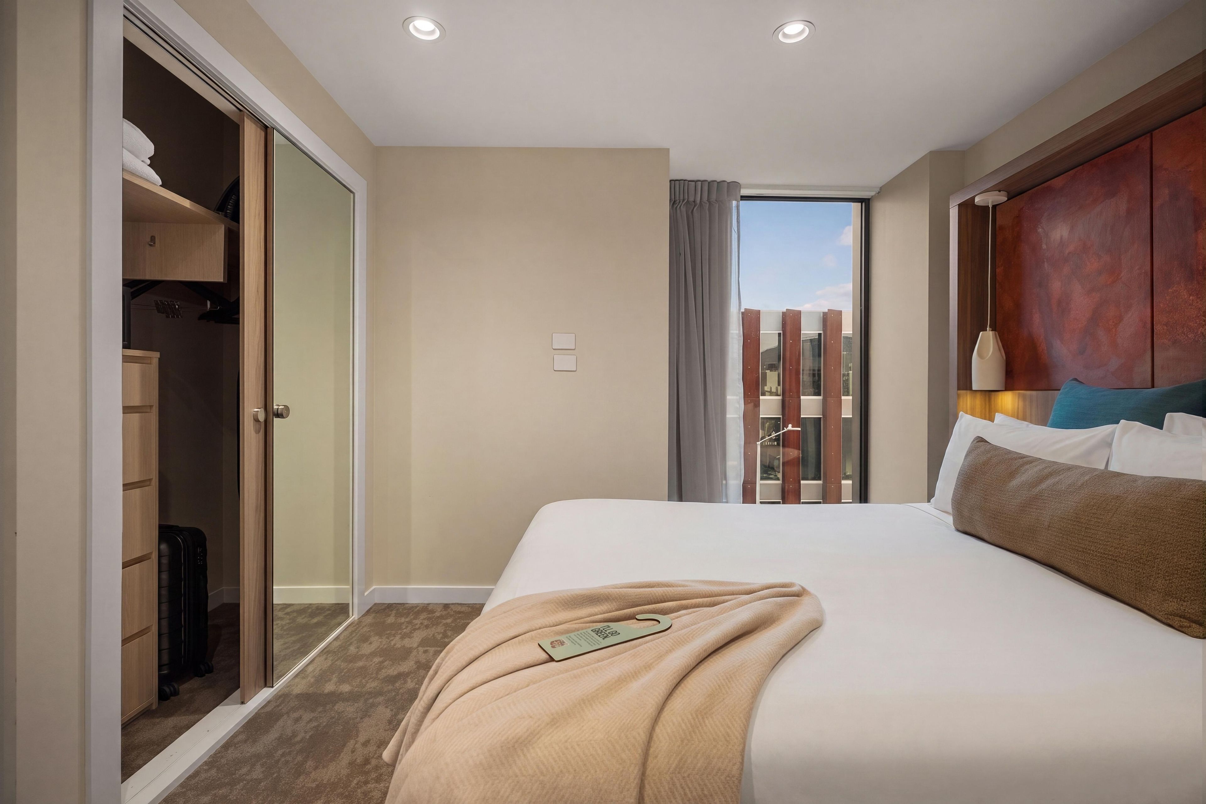 A hotel room with a bed, a wardrobe, a suitcase, and a view of the outside.