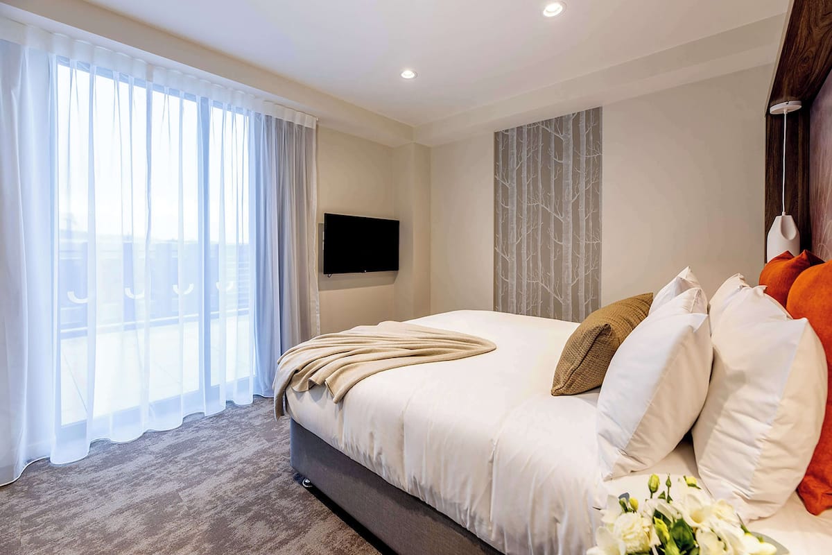 A modern hotel room with a large bed, a flat-screen TV mounted on the wall, and a view of the outdoors through sheer curtains.