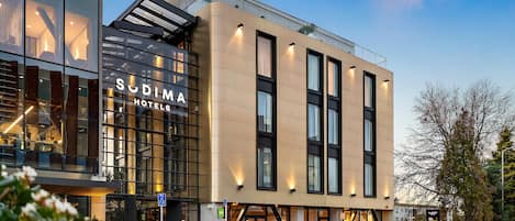 A modern hotel building with a glass facade, signage for 'Sudima Hotels', and a visible entrance.