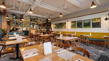 A restaurant interior with multiple dining tables, chairs, hanging pendant lights, a bar area, and a yellow bench along the wall.