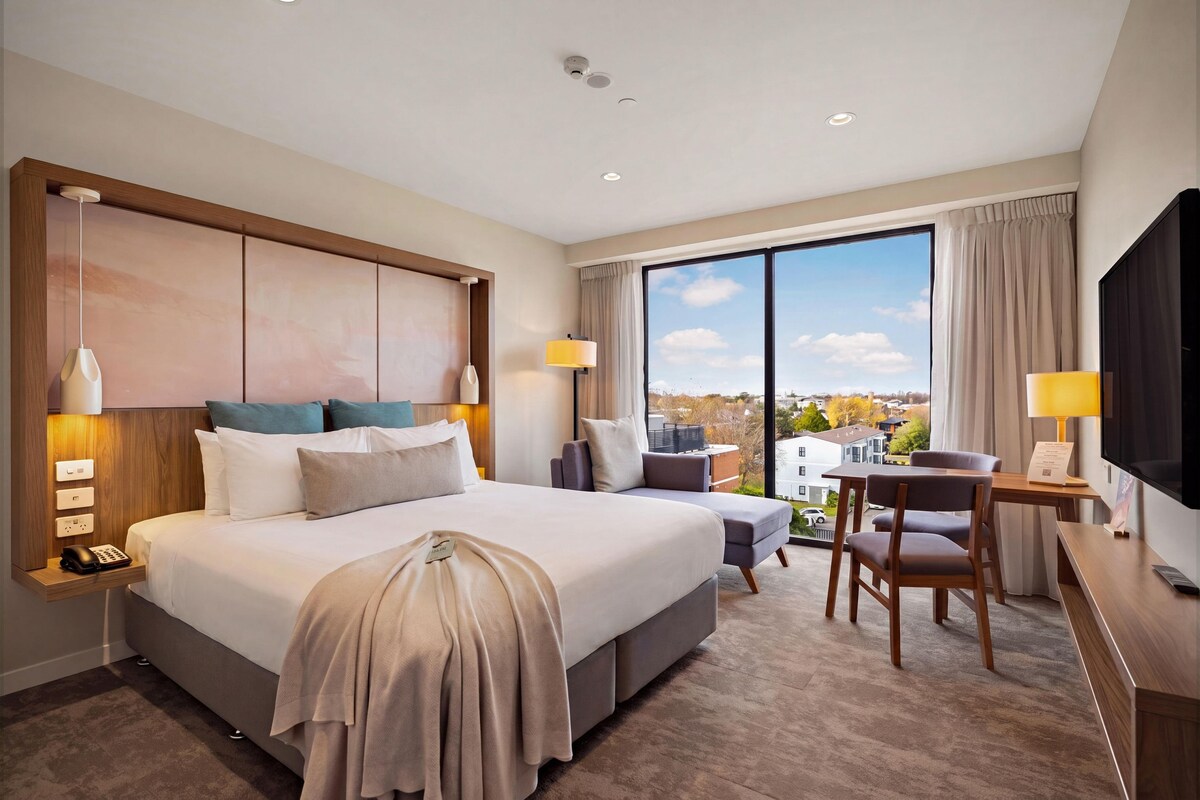 A hotel room with a large bed, bedside lamps, a desk with a chair, a television, and a view of the city.
