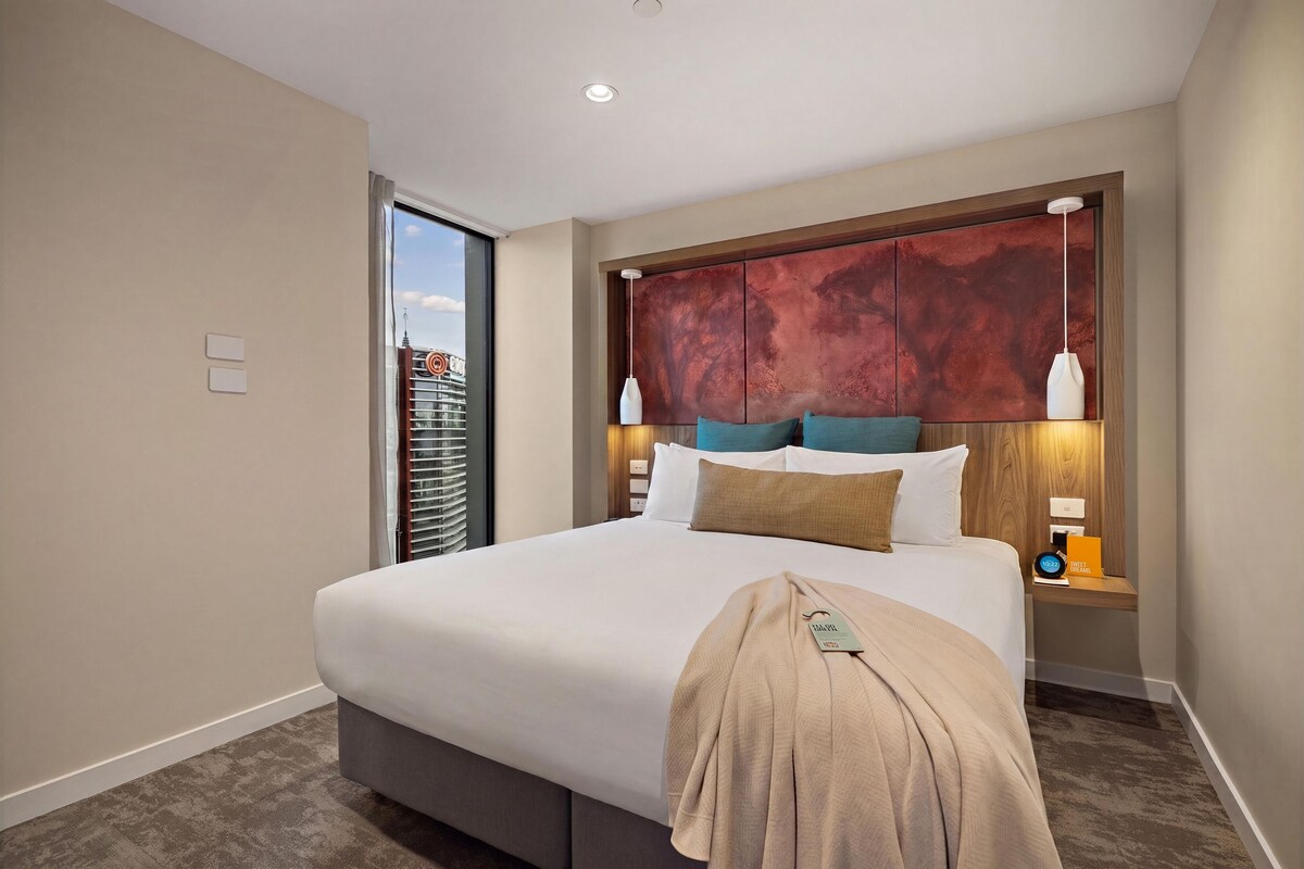 A hotel room with a large bed, a view of a cityscape through a window, and a modern headboard.