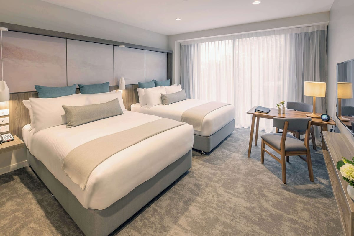 A hotel room with two beds, a desk, a chair, a lamp, and a TV.