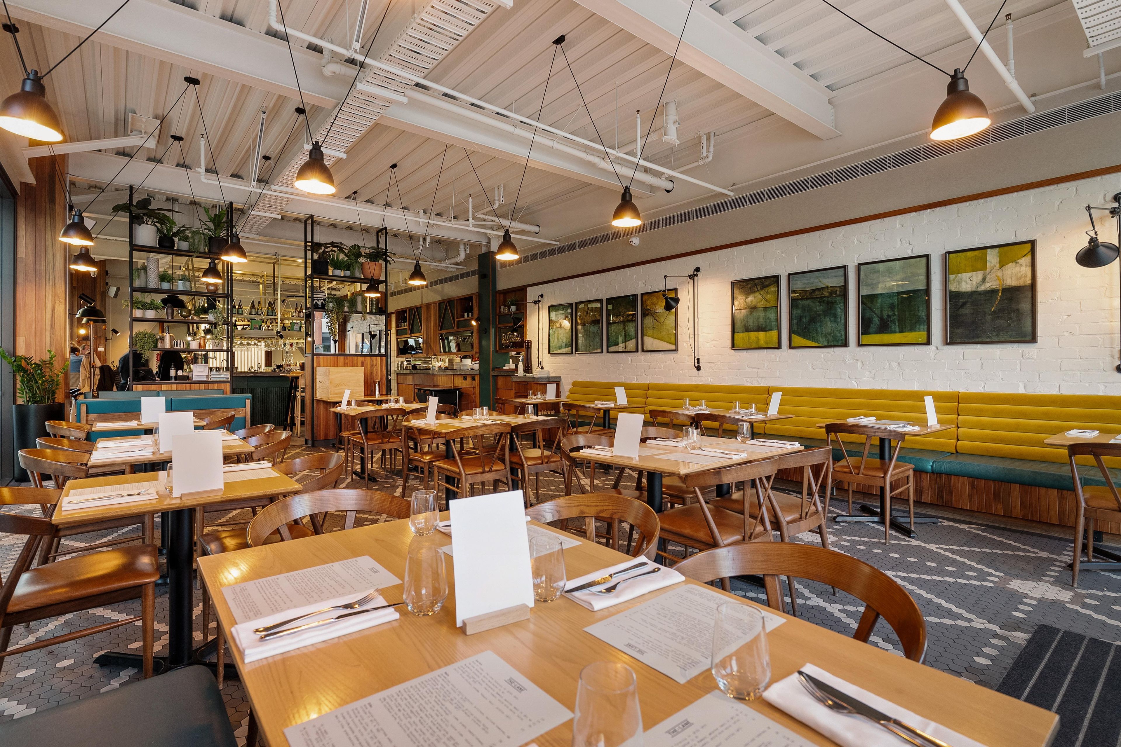 Ein Restaurantinneres mit mehreren Esstischen, Stühlen, hängenden Pendelleuchten, einer Bar und einer gelben Wandbank.