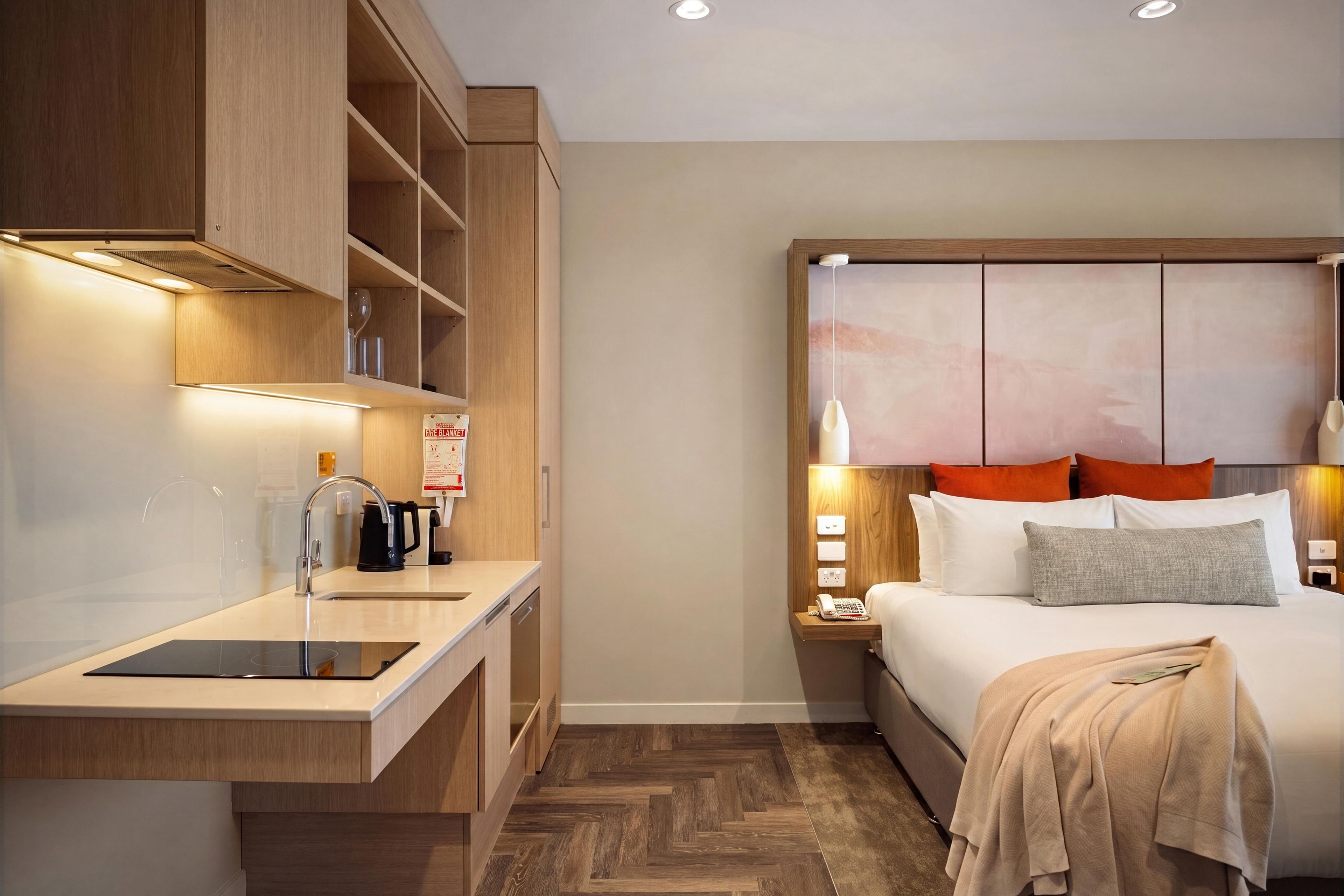 A hotel room with a wooden headboard, bedside table with a lamp, a bed with pillows, a nightstand, and a built-in kitchen area with a sink and coffee maker.