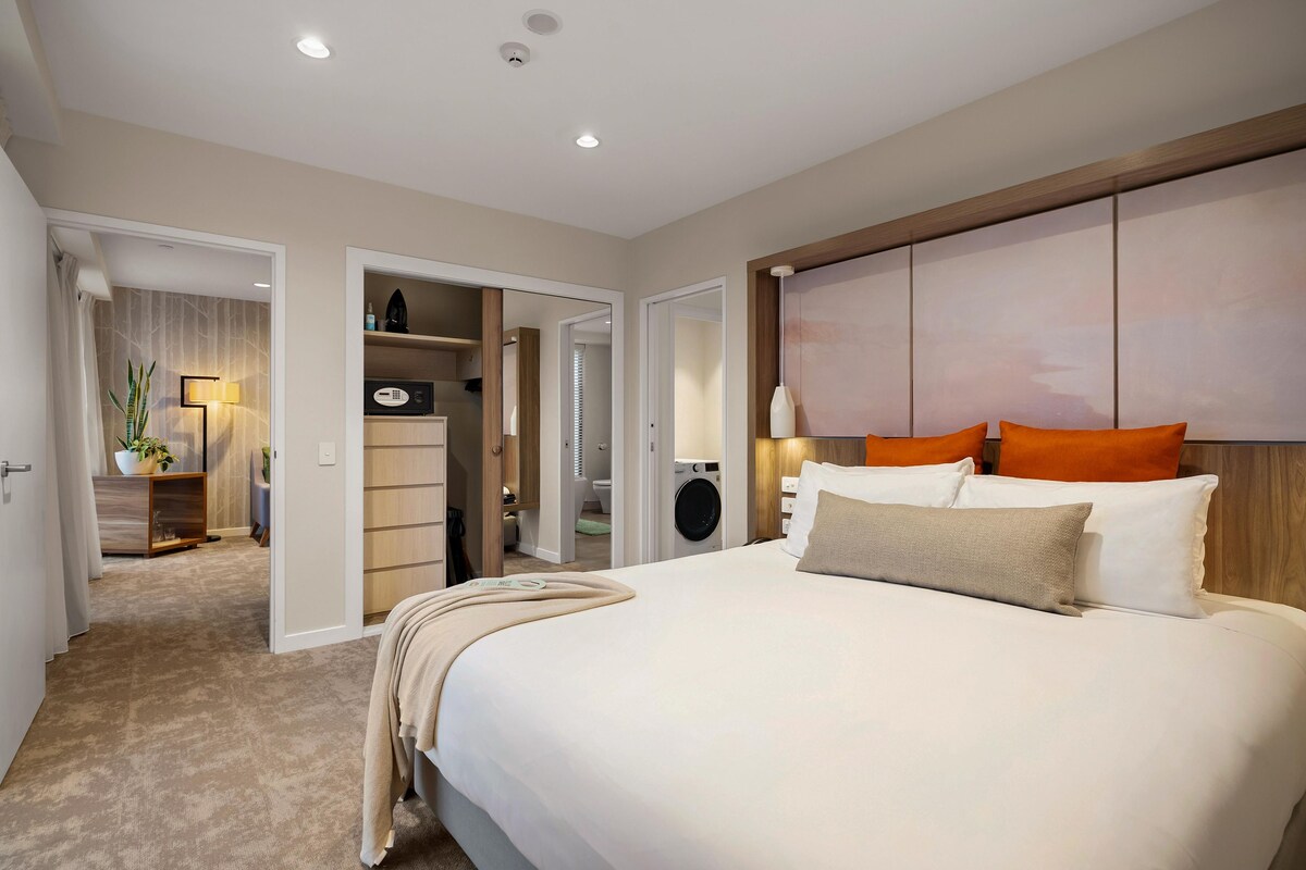A hotel room with a large bed, a wardrobe, a bathroom, and a hallway leading to another room.