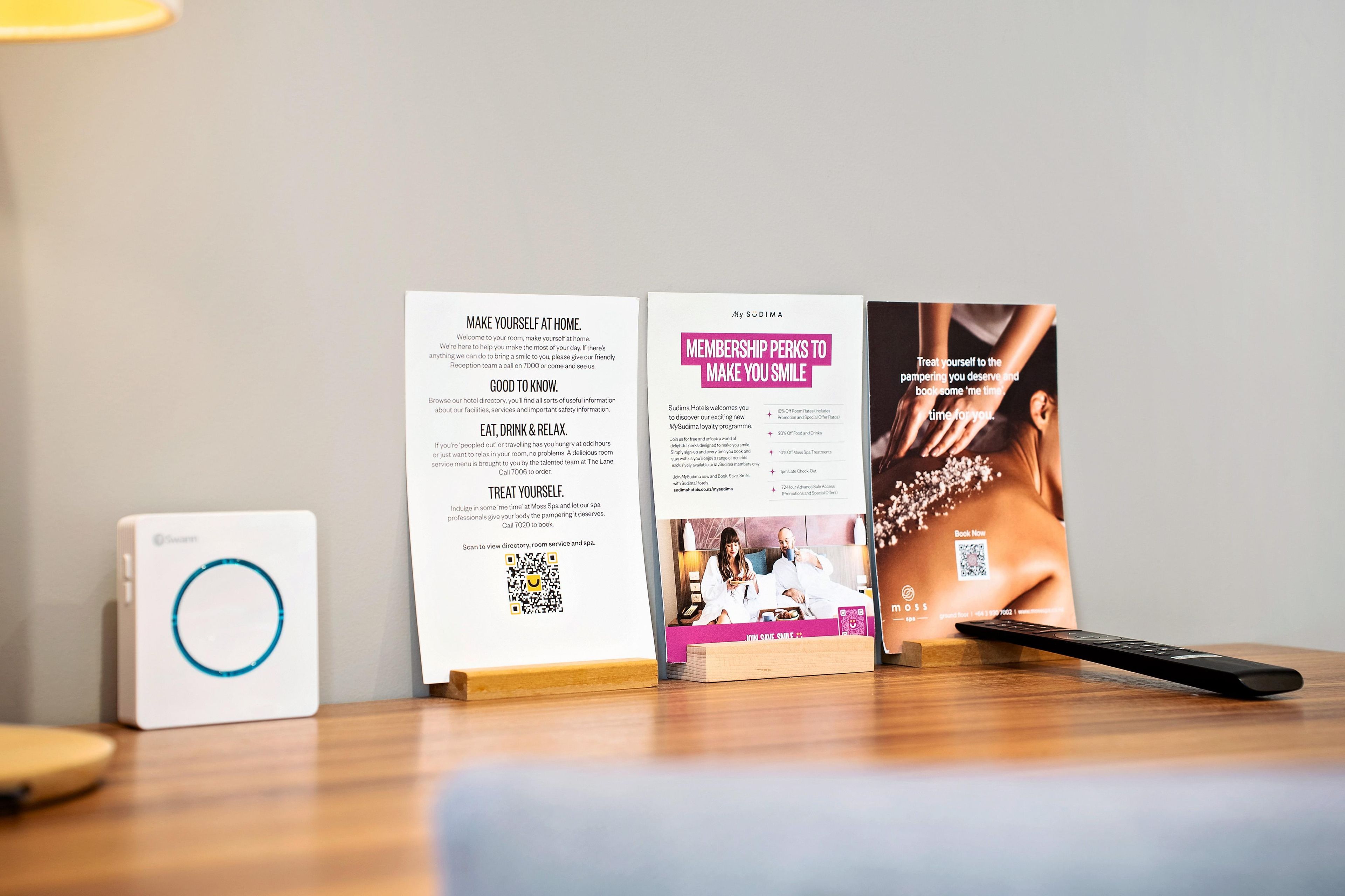 Un folleto con información sobre beneficios de membresía y servicios de spa, un código QR y un control remoto sobre una mesa de madera.