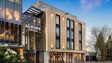 A modern hotel building with a glass facade, signage for 'Sudima Hotels', and a visible entrance.