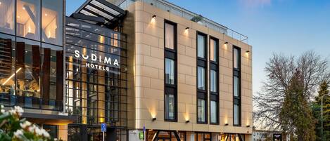 A modern hotel building with a glass facade, signage for 'Sudima Hotels', and a visible entrance.