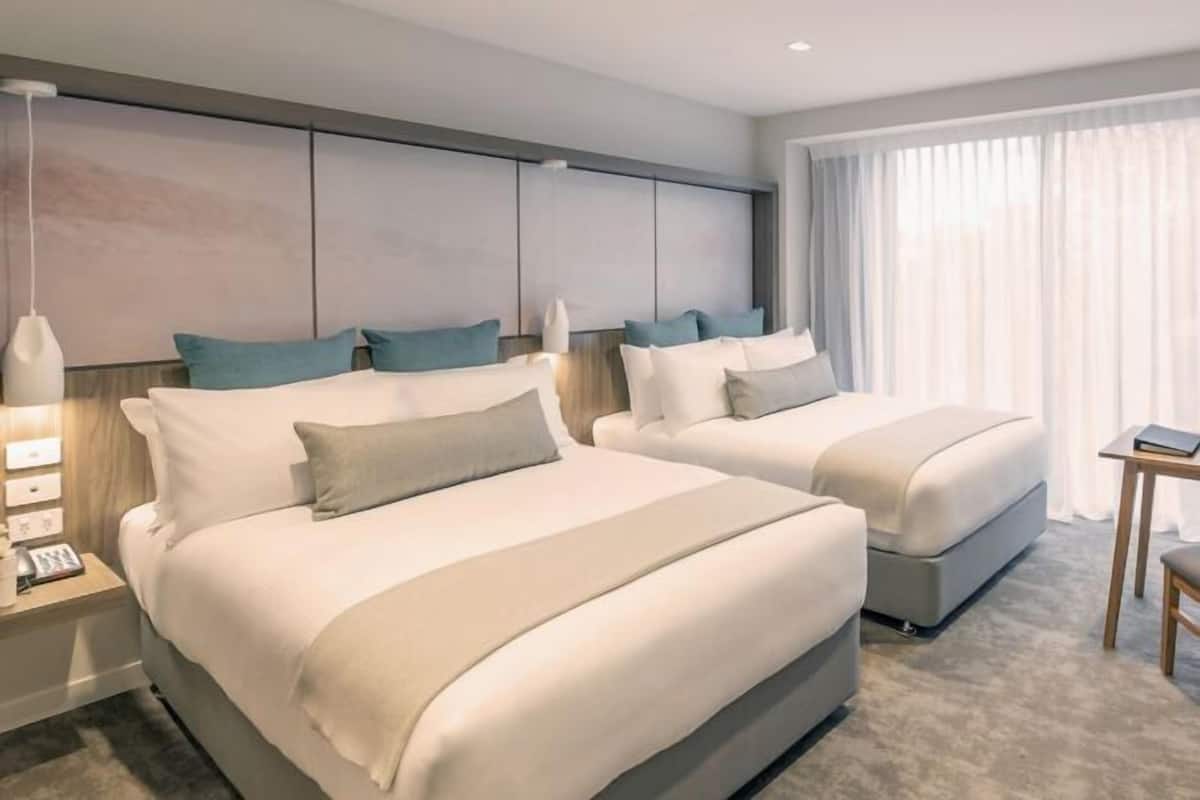 A hotel room with two beds, a bedside table with a lamp, a small table, and a window with curtains.