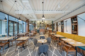 A modern restaurant interior with high ceilings, pendant lighting, and a mix of wooden and upholstered furniture.