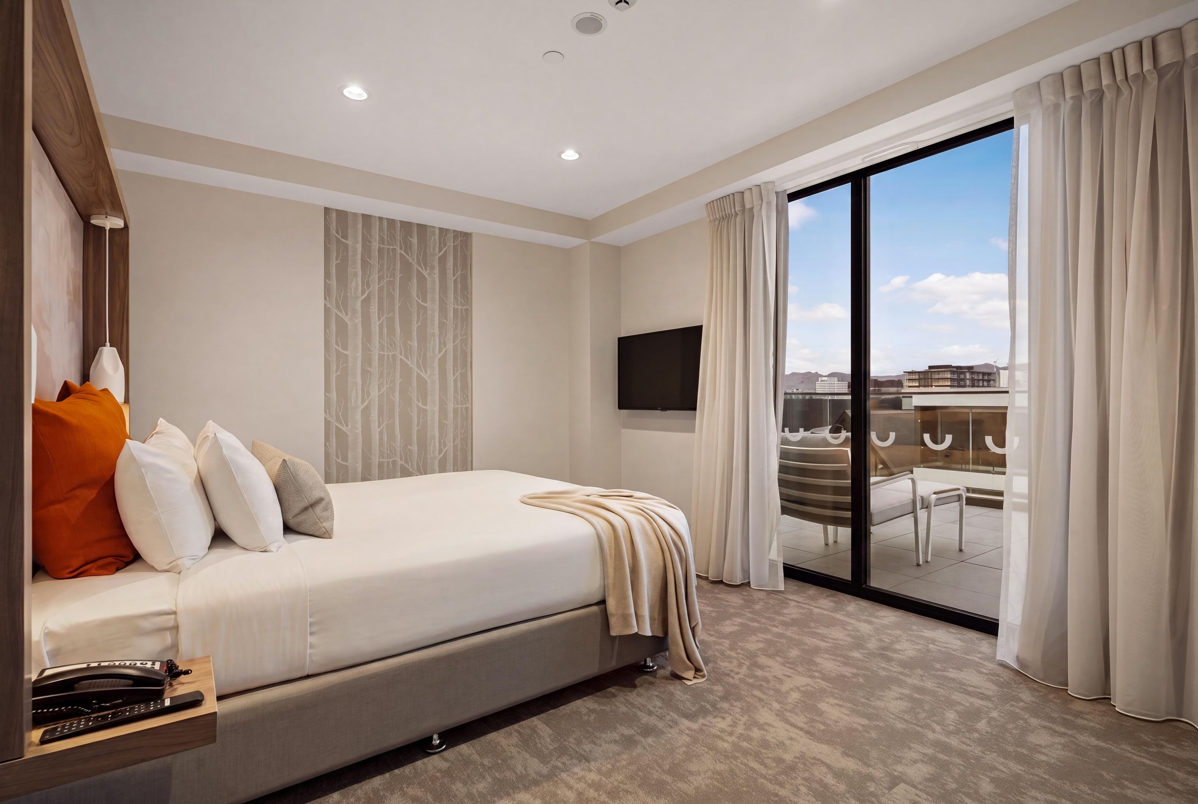 A hotel room with a bed, a desk with a telephone, a flat-screen TV, and a sliding glass door leading to a balcony.