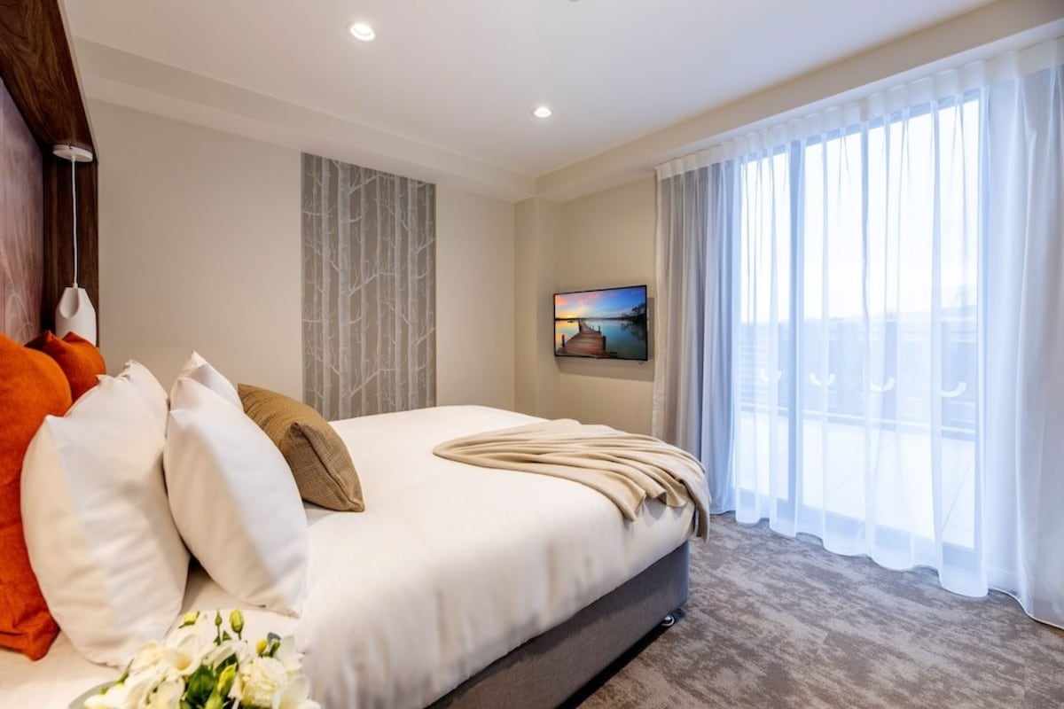 Una habitación de hotel moderna con una cama grande, un televisor y vistas de la ciudad a través de cortinas translúcidas.