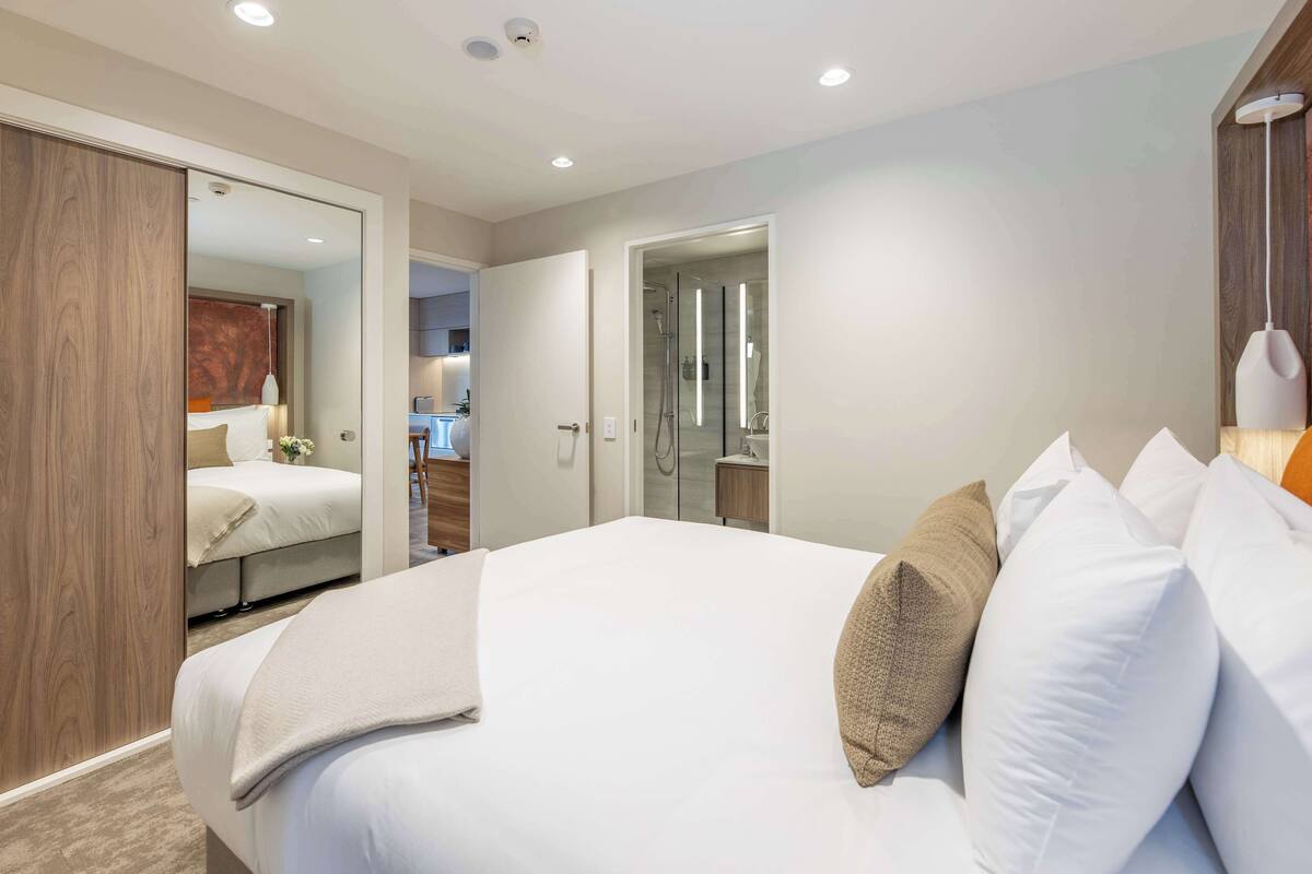 A modern hotel room with a large bed, a walk-in closet, a bathroom with a glass shower, and a small desk area.