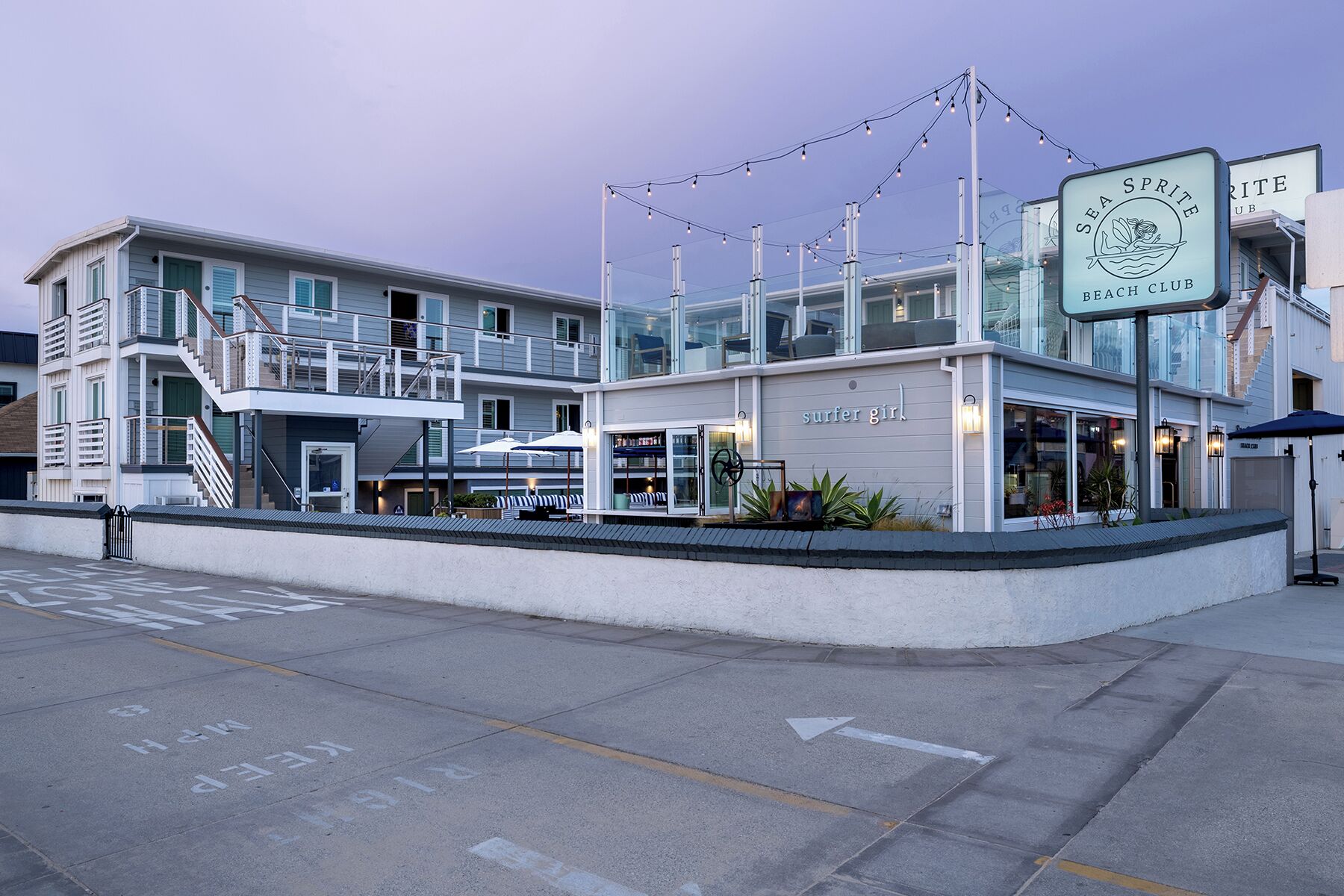 A beachfront building with a 'Surfer Girl' sign, a balcony, and a beach club sign.