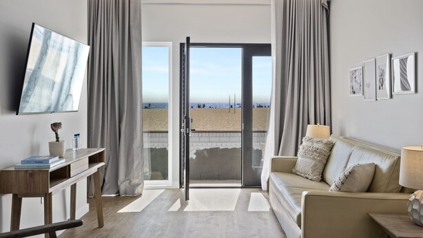 A modern living room with a sofa, a coffee table, a TV, and a desk. There is a large window with curtains, and a view of the beach is visible through the glass door. - Sea Sprite Beach Club (Hermosa Beach)