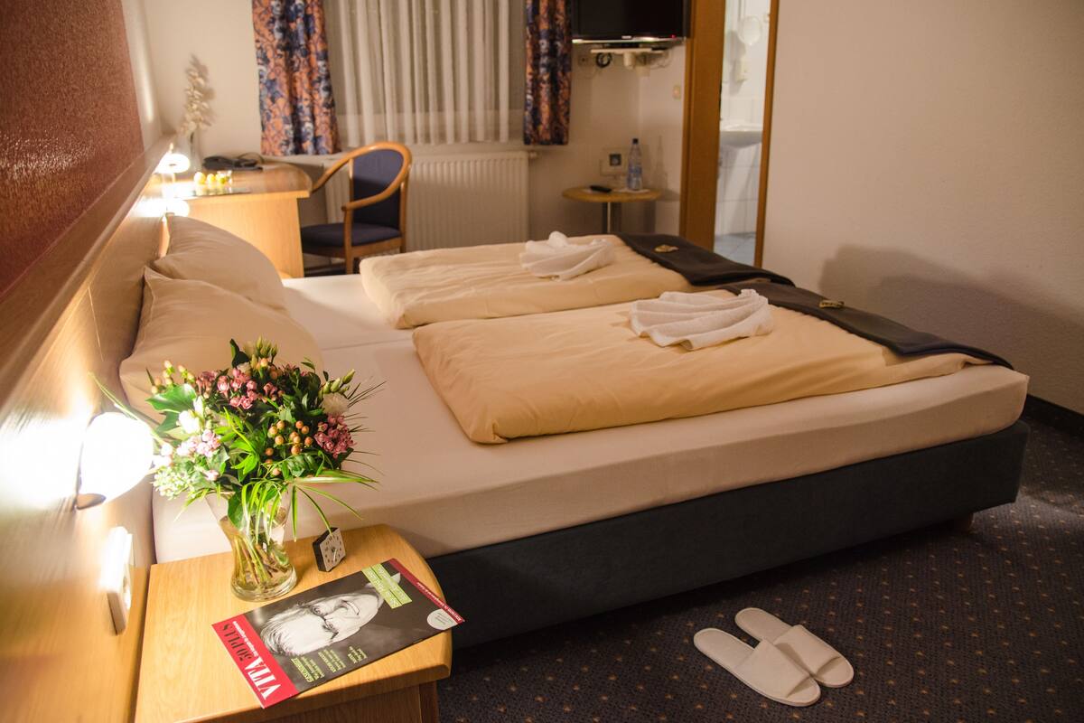A hotel room with two beds, a desk with a lamp, a small table with a vase of flowers, a book, and slippers on the floor.