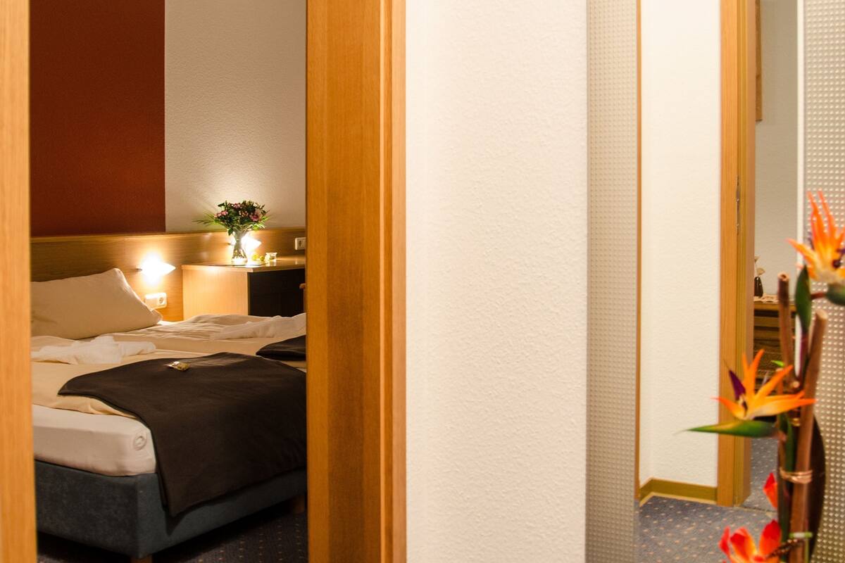 A hotel room with a bed, bedside table, lamp, mirror, and a potted plant.