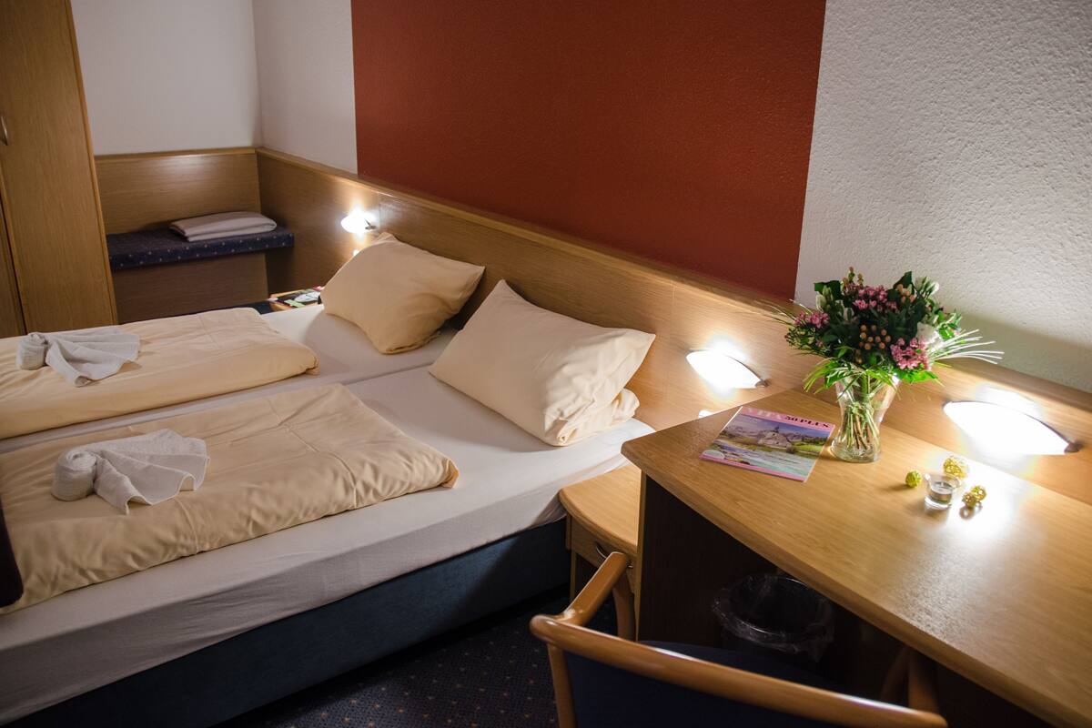 A hotel room with a bed, a desk, a chair, a trash bin, and a vase with flowers.